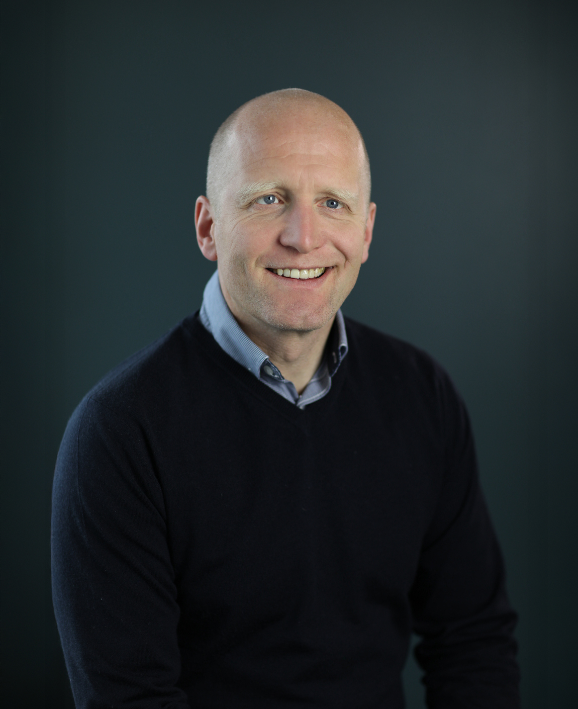 Professional portrait of smiling bald man in dark sweater and light blue collared shirt against dark teal background.