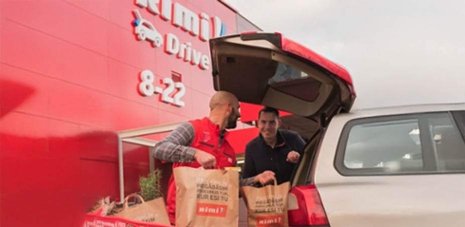 Two Rimi employees loading grocery bags into customer's car trunk at drive-through service station with red Rimi Drive signage showing 8-22 hours