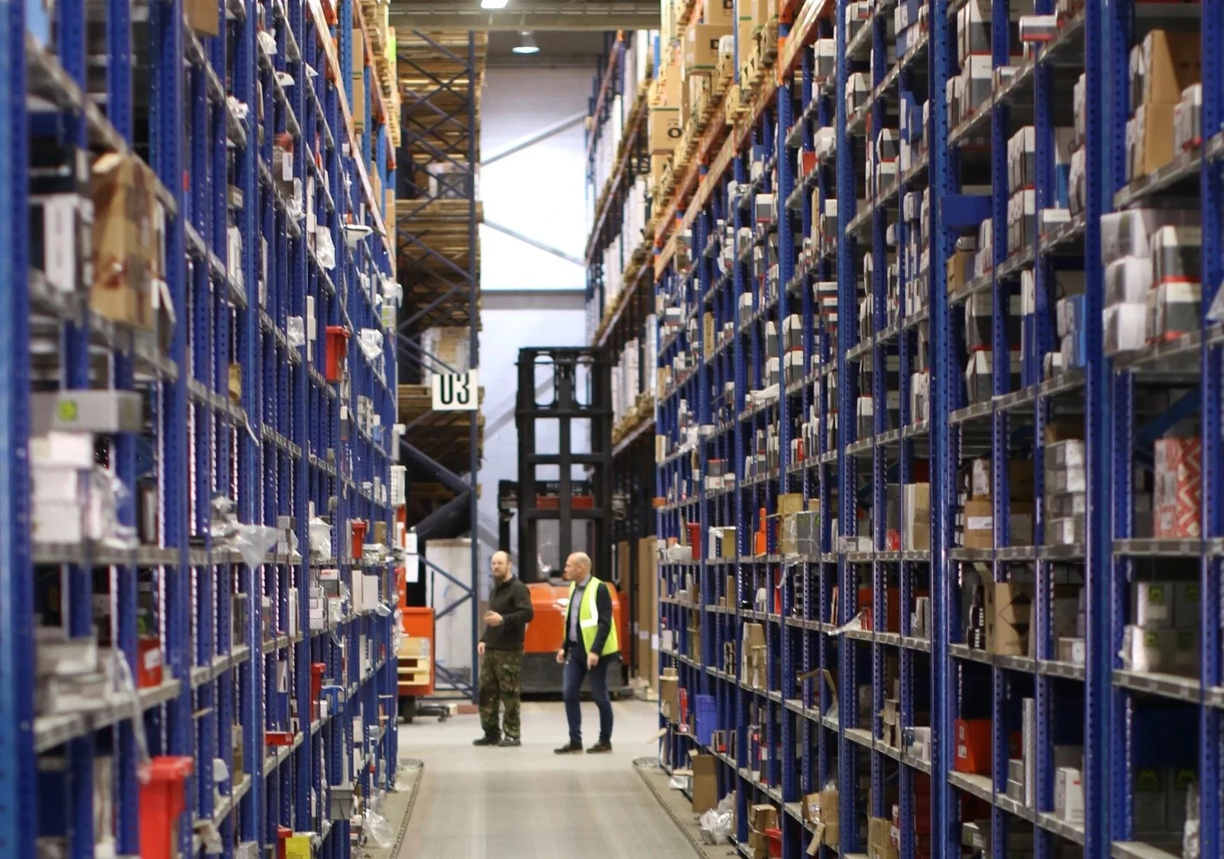 Two workers standing in a large warehouse aisle between tall blue shelving units filled with boxes, with a forklift visible.