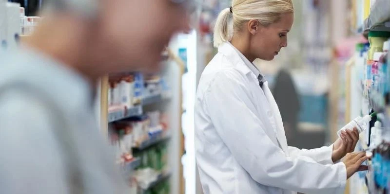 Pharmacy worker in a white lab coat reading a product label while standing in a medication aisle with shelves of products.