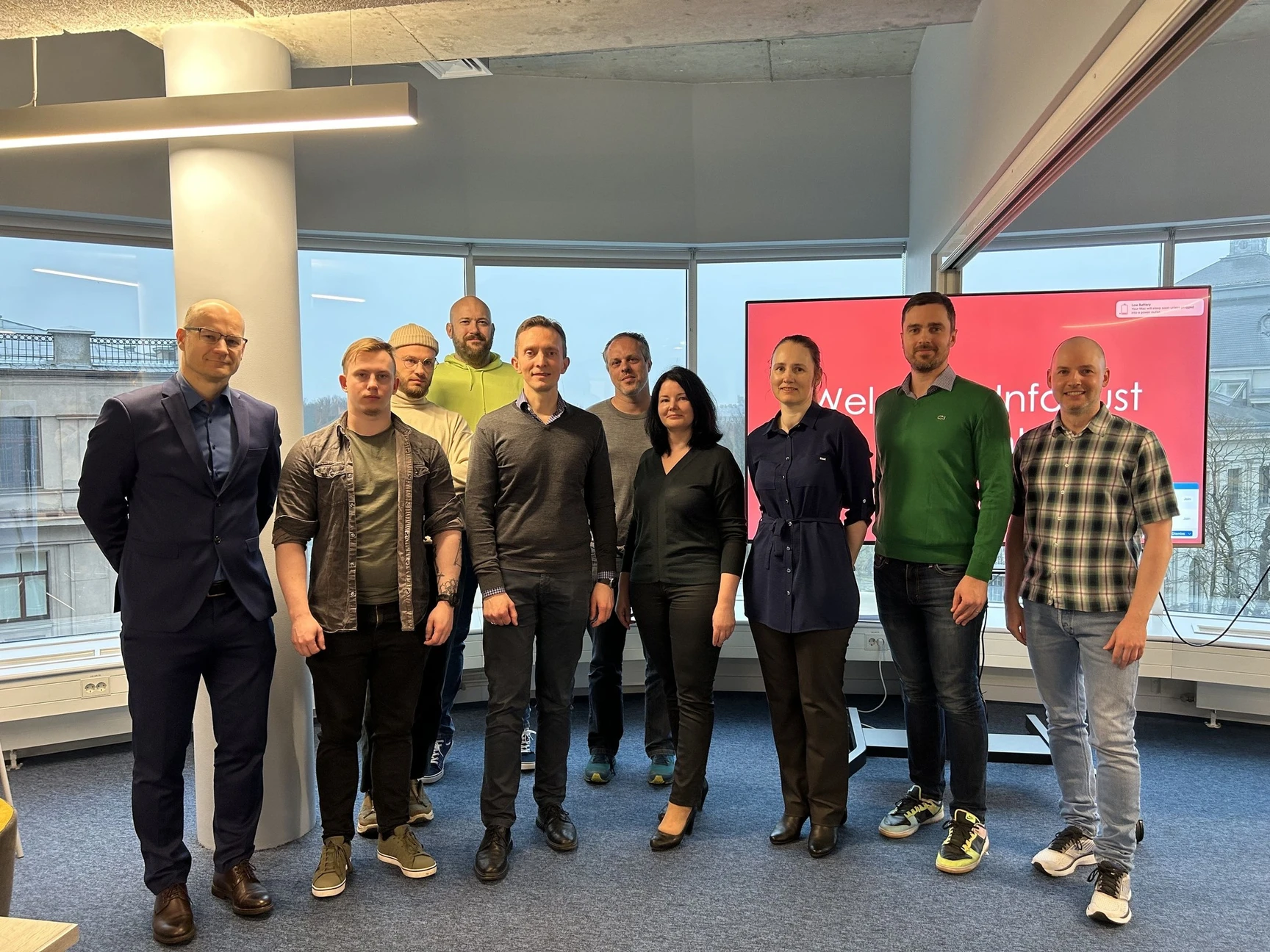 A group of ten people posing for a photo in a modern office with large windows and a red screen in the background.