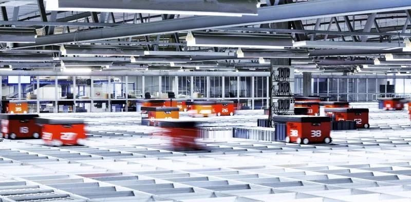 Automated warehouse with multiple numbered red robots moving across a large grid floor under bright ceiling lights.