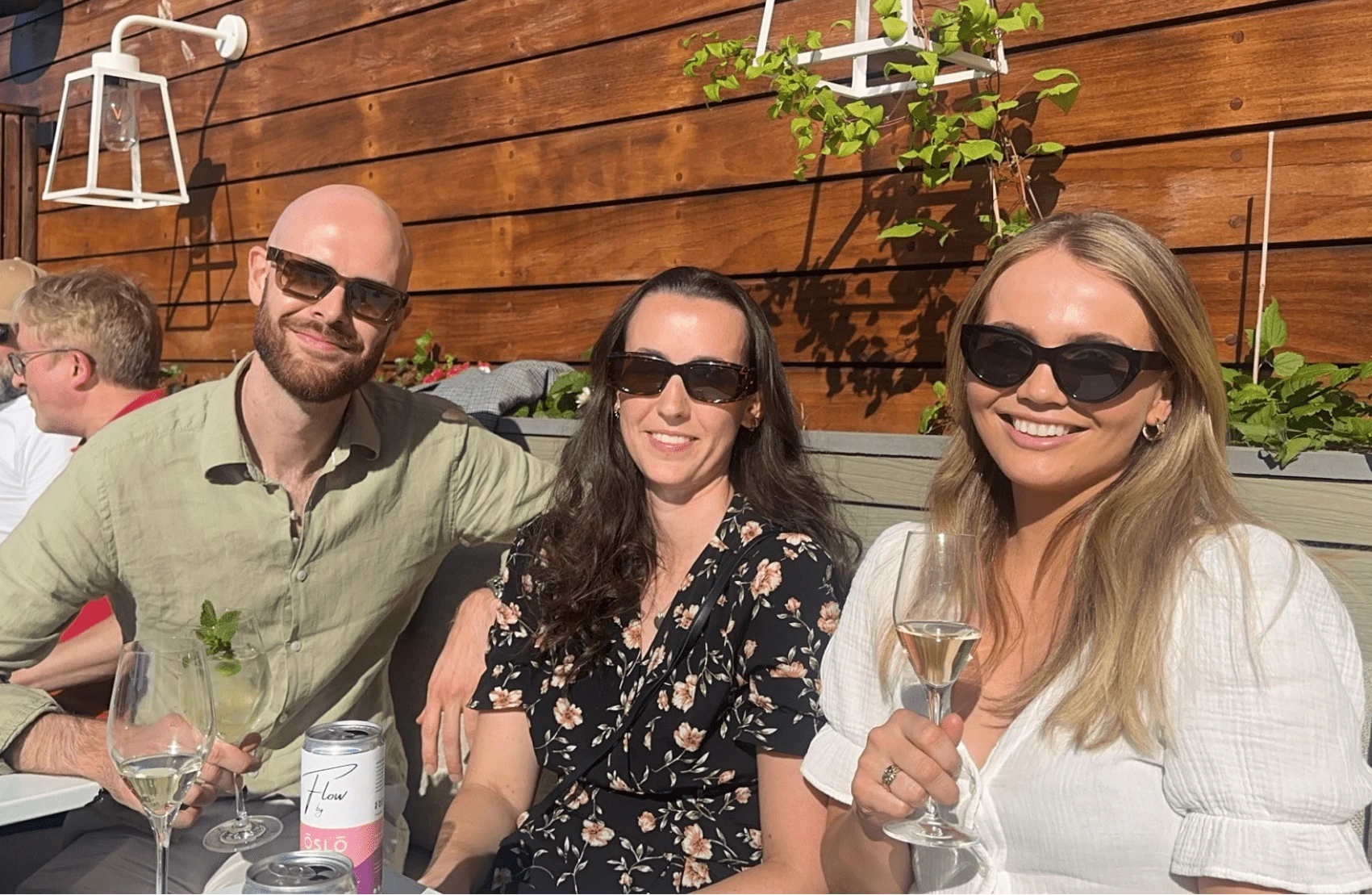 Three smiling people with sunglasses sitting outdoors in the sun with drinks, in front of a light wooden wall.