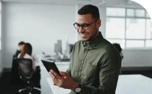 Smiling man in glasses and green shirt looking at a tablet in a modern office environment with colleagues in the background.