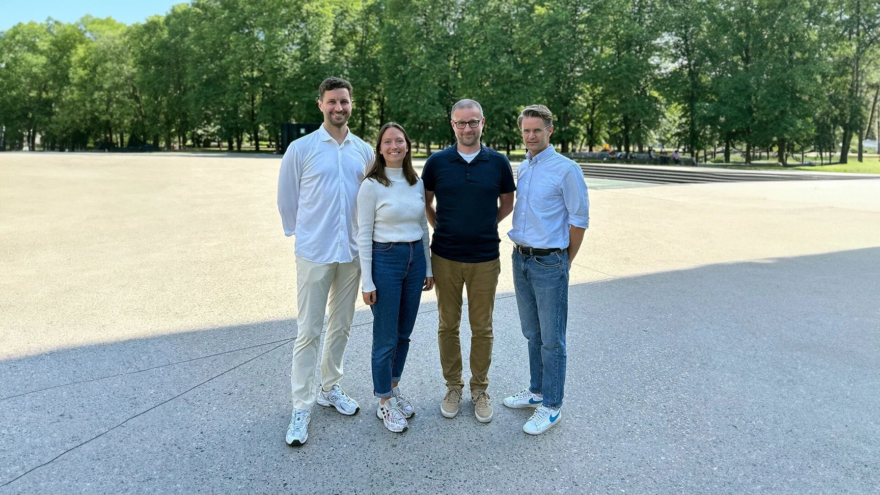 Four colleagues standing together outdoors on a sunny day in a park setting with trees in the background.
