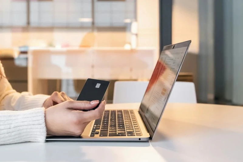 Laptop on a table with a person sitting in front with a credit card in its hands.