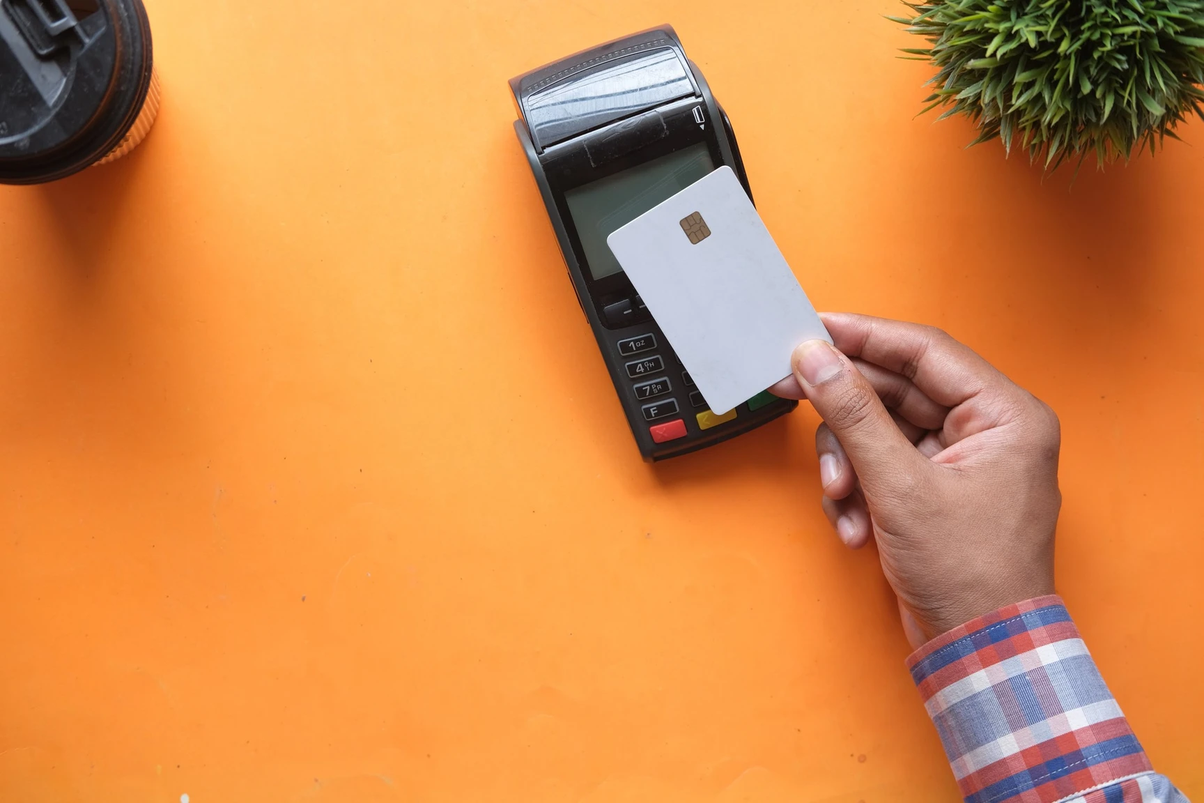 Person holding a white chip card to a card payment terminal on an orange background, with a coffee cup and plant nearby.