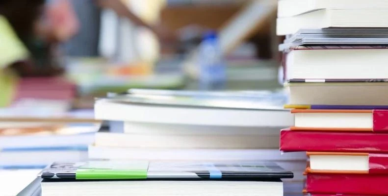 Several stacks of books with colorful spines arranged on a table in a bright indoor setting with a blurred background.