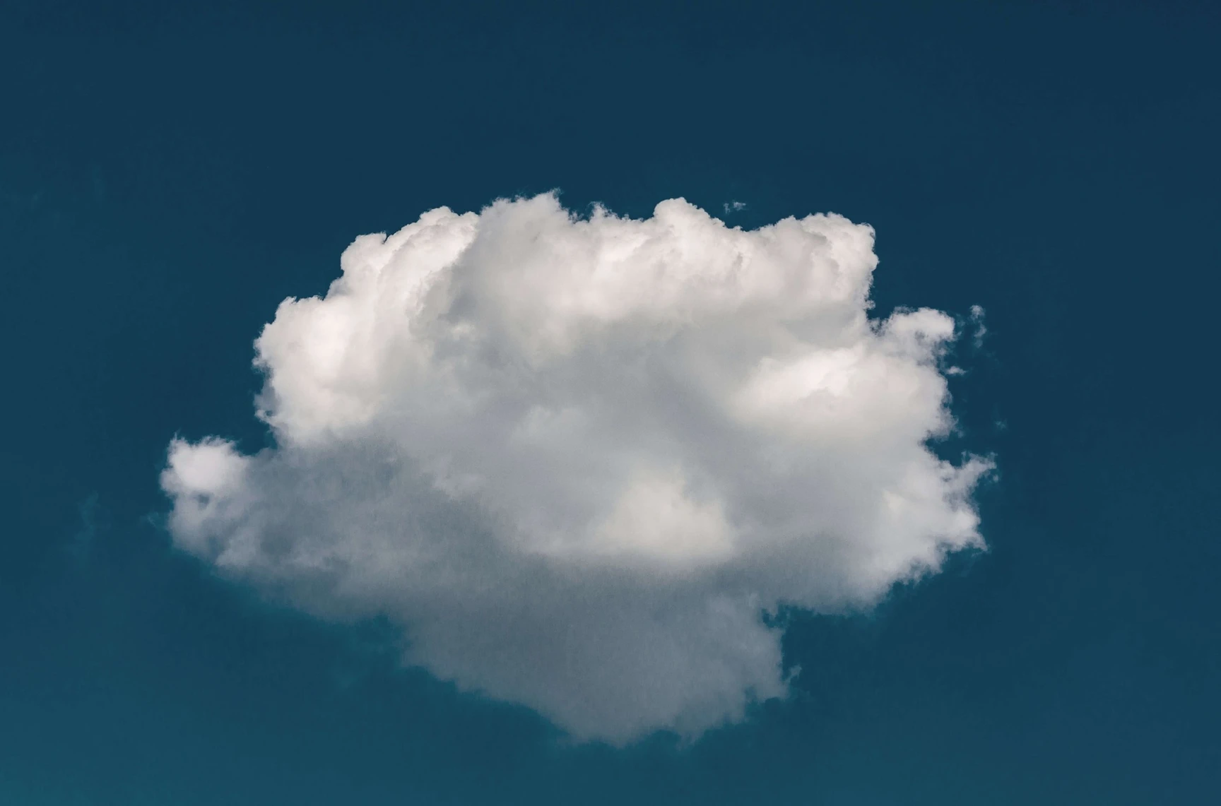 Close-up photograph of a single large white cumulus cloud set against a deep teal blue sky with clear visibility.