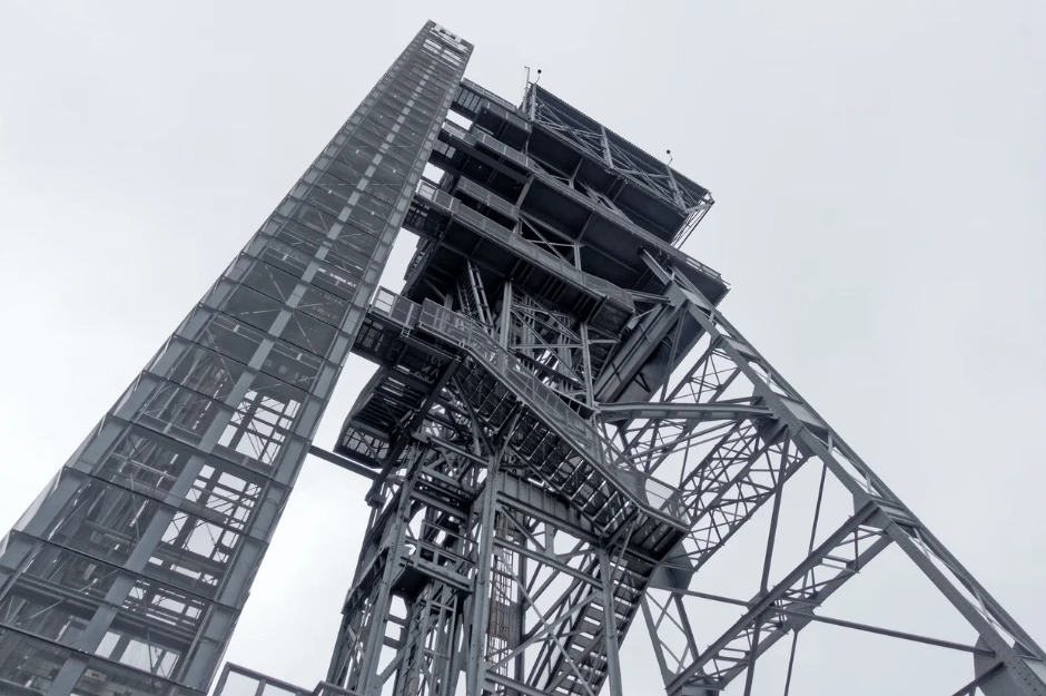 Low-angle view looking up at a large industrial steel framework tower and structure against an overcast grey sky.