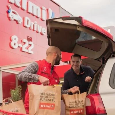 Rimi Drive store employee in a red vest loading branded grocery bags into a customer's car boot outside a store.