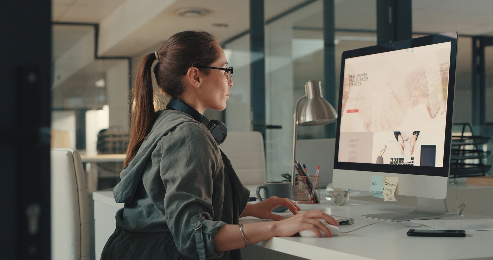 Woman with glasses and headphones around her neck working at a desktop computer in a modern office environment.