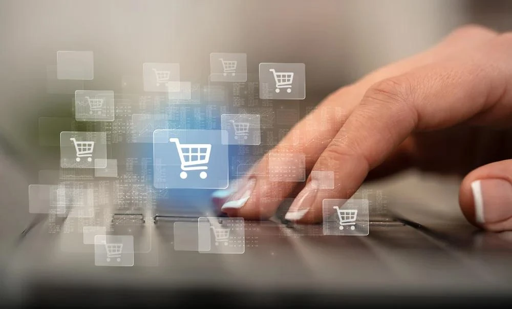 Hand pressing a laptop touchpad surrounded by floating digital shopping cart icons on a light background.