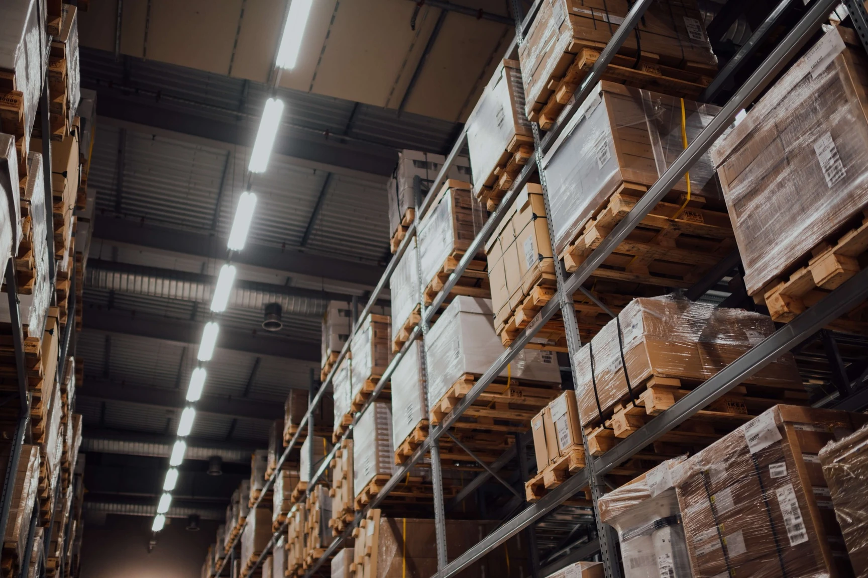 Tall warehouse shelving units stacked with wrapped pallets and cardboard boxes under fluorescent ceiling lights.