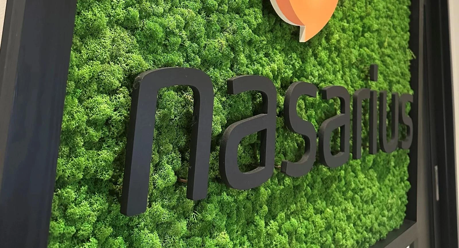 Nærbilde av en mosevegg med firmanavnet Nasarius i svarte bokstaver og en del av en oransje logo øverst.